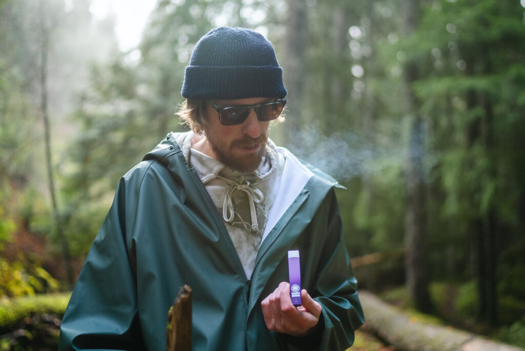 The image shows a man in a green jacket vaping cannabis in the forest.